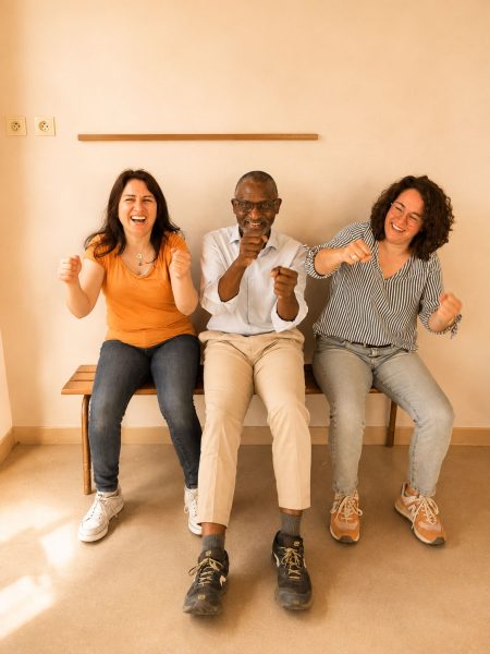 Trois adultes assis rient ensemble en mimant un geste avec leurs mains